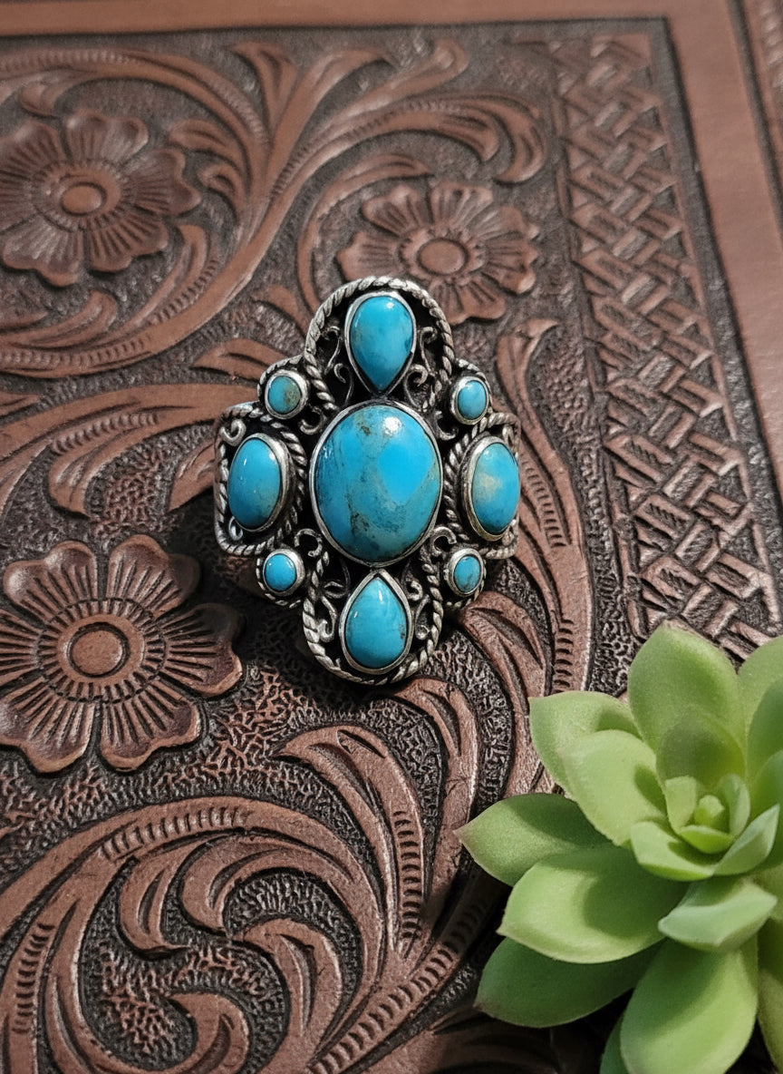 Sterling silver ring with ornate filigree and multi-stone turquoise cluster in oval and teardrop shapes, shown on tooled leather background