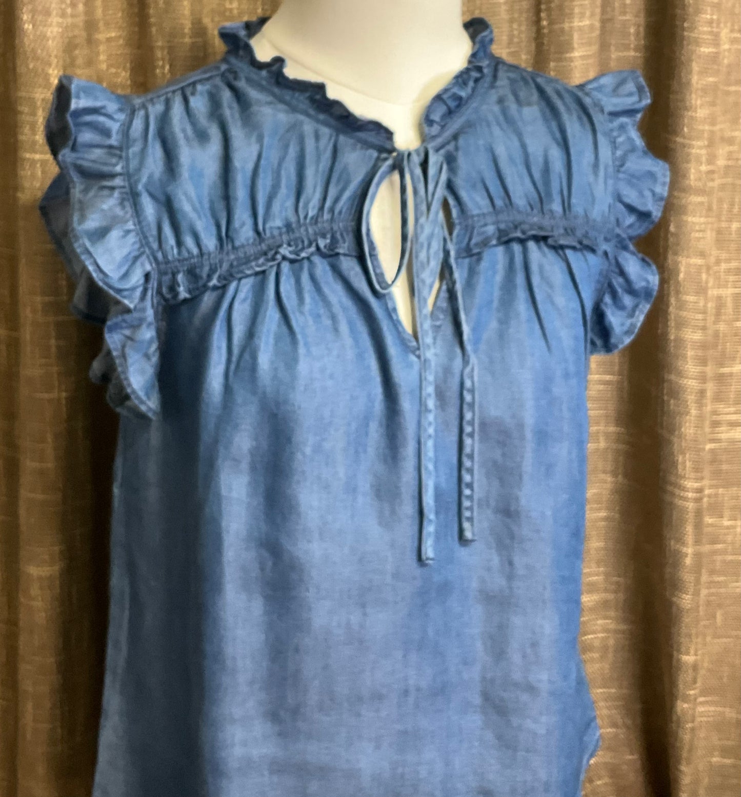 Jenna Chambray Blouse in soft blue lyocell—sleeveless top with high neckline, feminine neck tie, keyhole front detail, ruffle trim, and gently ruched yoke for subtle texture and charm.