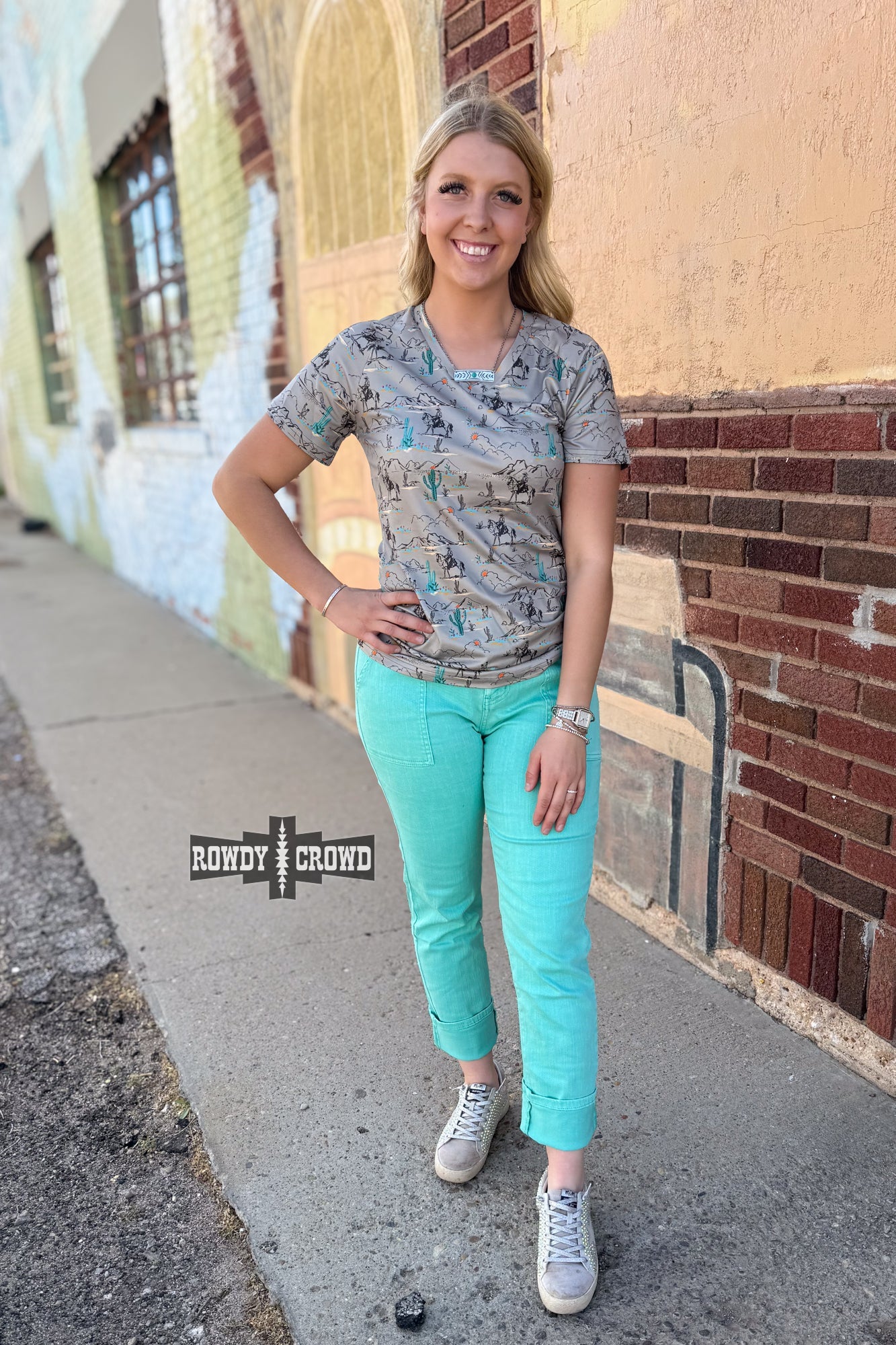 Blooming Desert Top featuring a detailed desert landscape print with cowboys on horseback, vibrant cacti, and blooming wildflowers. Soft, breathable fabric with a relaxed fit. Available in sizes Small-3XL.