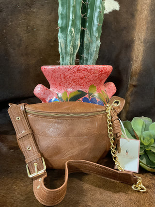 A brown cognac vegan leather sling bum bag with an adjustable strap, displayed in front of a red vase with a green cactus in the background.