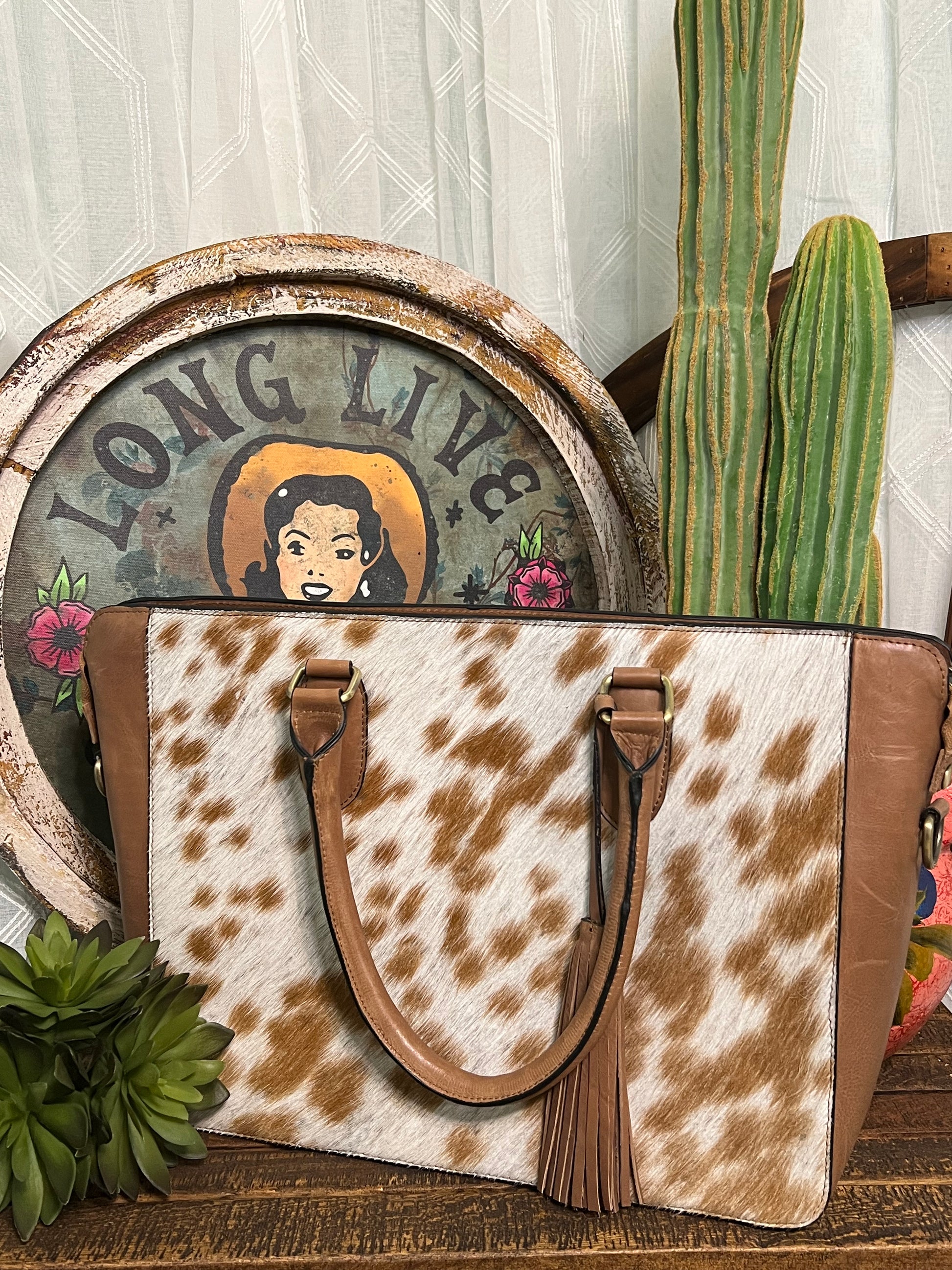 Brushy Creek Cowhide Tote:  handcrafted leather and cowhide tote purse, featuring a light brown and white cowhide front panel, a tassel on the front strap, a zippered closure, and an optional longer carrying strap. Approximate size: 14" x 10.5".