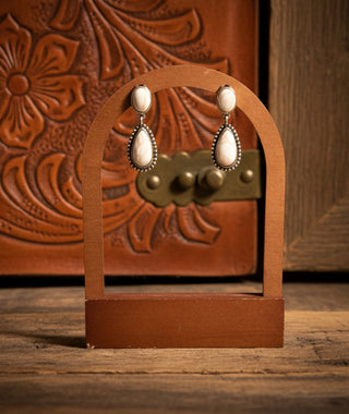 A pair of western-style drop earrings with white teardrop-shaped stones and silver-toned beaded borders, displayed on a small wooden stand against a brown embossed leather background.