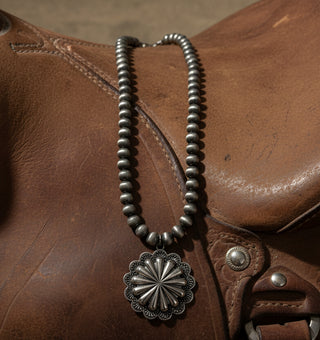 Silver-tone beaded necklace with a large engraved concho pendant resting against brown leather.