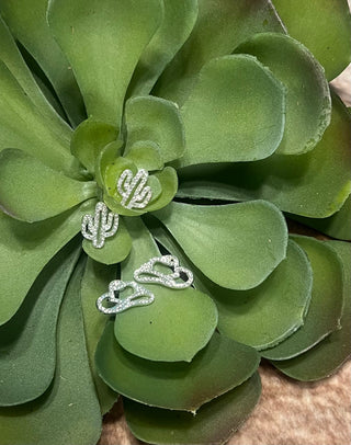 The image features a pair of Disco Cowgirl Earrings displayed on a green plant. The earrings are designed either as a cowboy hat or a cactus, sparkling with rhinestone pave.