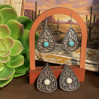 Two pairs of teardrop earrings with ornate western scrollwork, one silver toned with a turquoise center stone and one copper toned with a white center stone, displayed on a wooden stand with rustic western décor.
