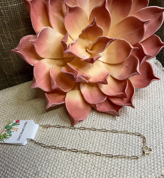 A gold-plated necklace with a paperclip chain design and a heart-shaped front lobster clasp with CZ accents, displayed on a textured background with a large pink flower.