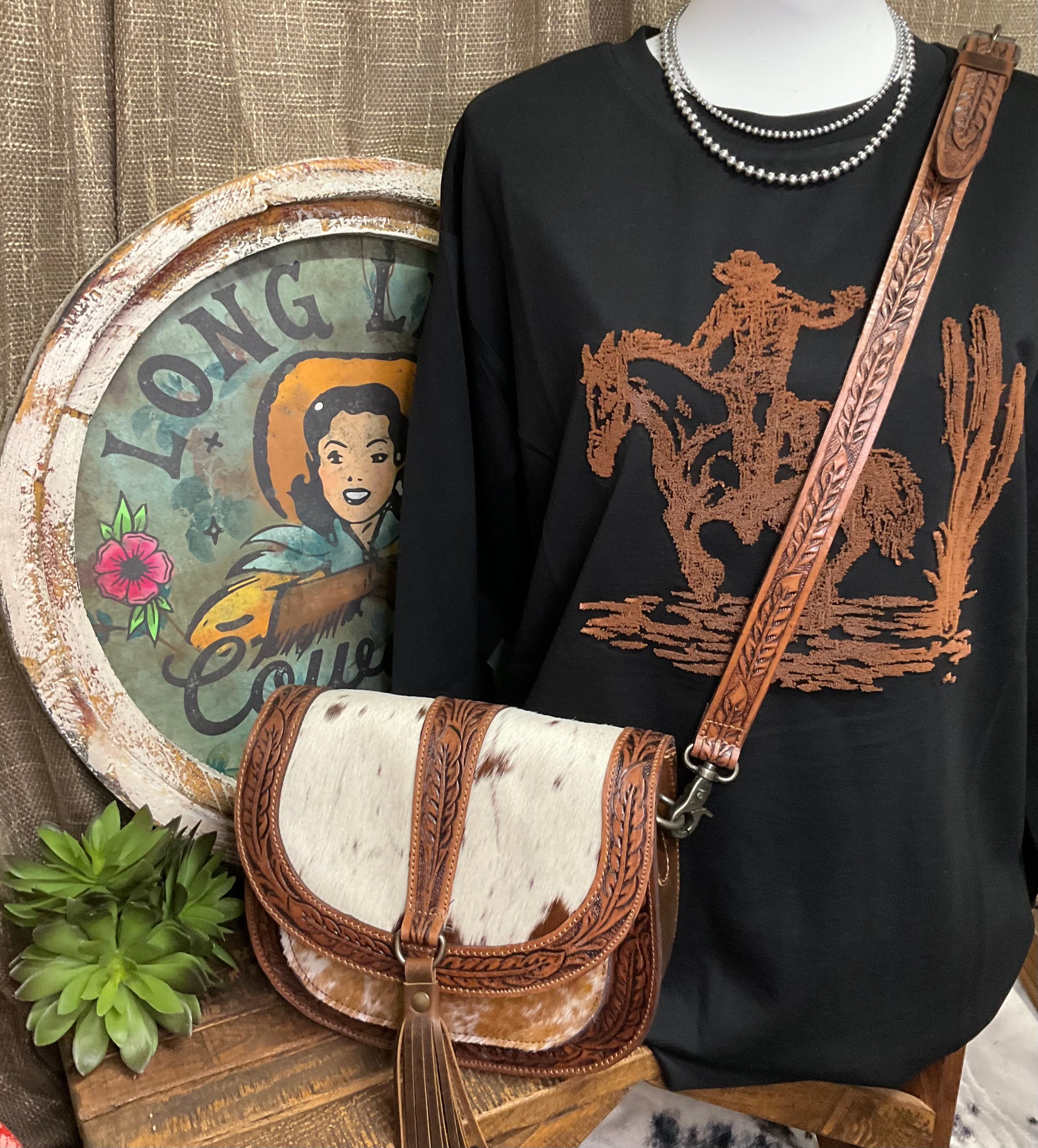 A stylish crossbody bag called the Paris Cowhide & Tooled Crossbody. The bag features a unique combination of genuine cowhide and intricately tooled leather accents. It has a structured shape with a secure flap closure, an adjustable leather strap, and a compact size perfect for essentials. The natural cowhide pattern adds a rustic charm, making it a standout accessory for both casual and dressier outfits. Ideal for those who love Western-inspired style with a touch of elegance.