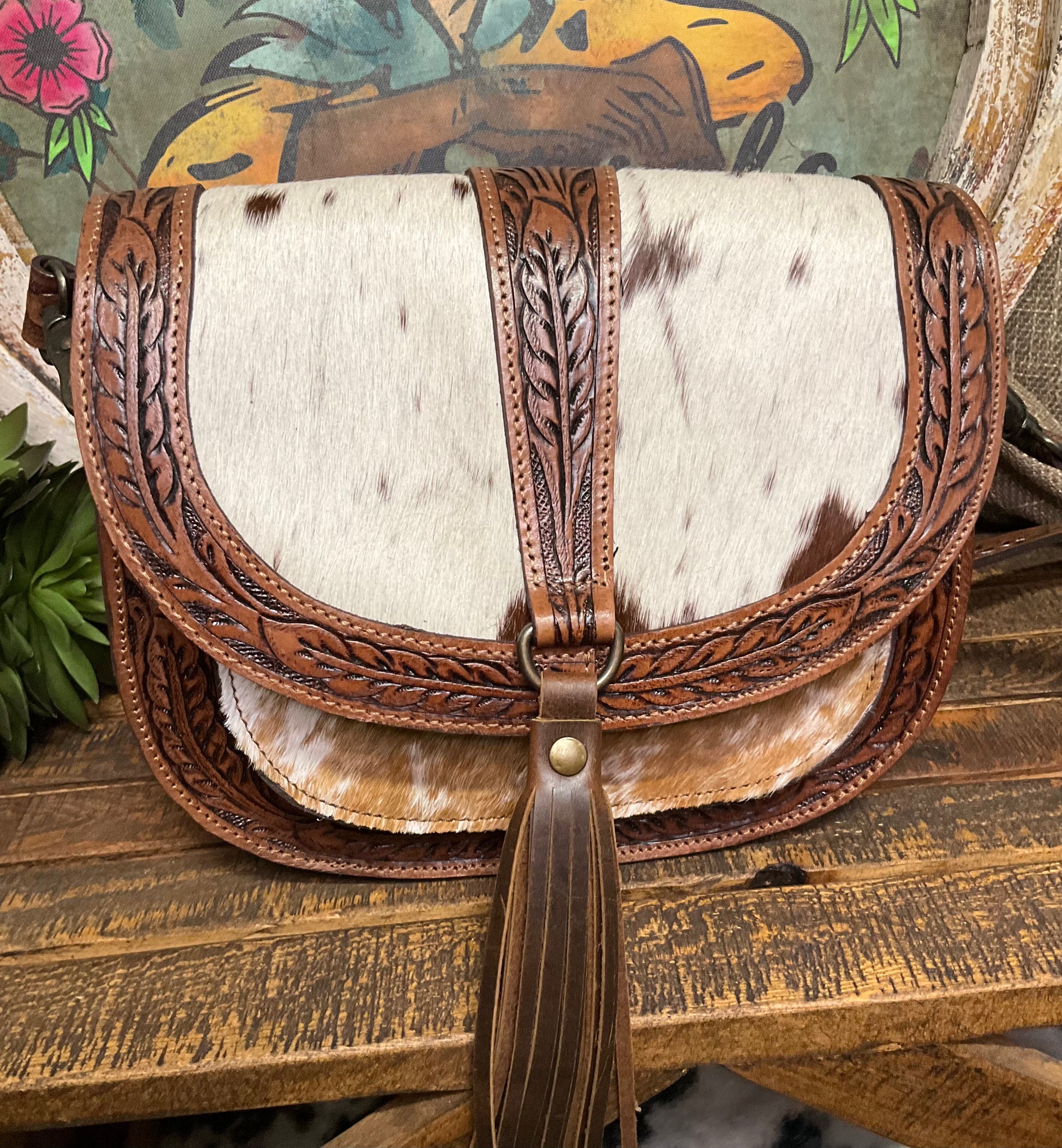 A stylish crossbody bag called the Paris Cowhide & Tooled Crossbody. The bag features a unique combination of genuine cowhide and intricately tooled leather accents. It has a structured shape with a secure flap closure, an adjustable leather strap, and a compact size perfect for essentials. The natural cowhide pattern adds a rustic charm, making it a standout accessory for both casual and dressier outfits. Ideal for those who love Western-inspired style with a touch of elegance.