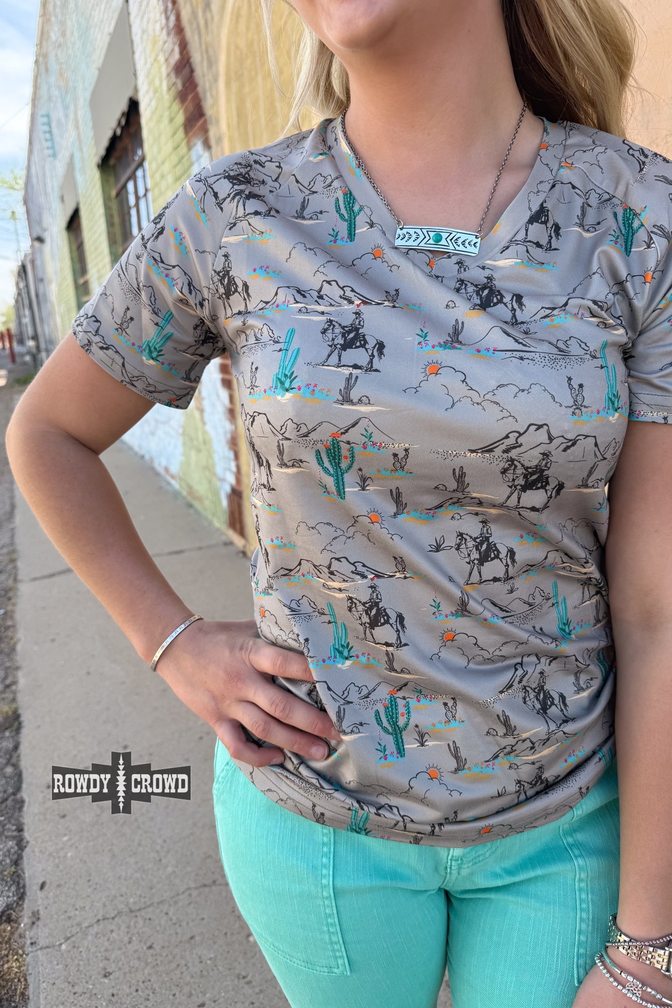 Blooming Desert Top featuring a detailed desert landscape print with cowboys on horseback, vibrant cacti, and blooming wildflowers. Soft, breathable fabric with a relaxed fit. Available in sizes Small-3XL.