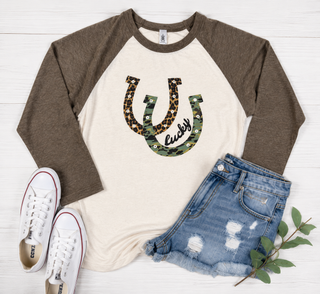 Luck Runs Deep cream and army green raglan tee with leopard and camo Lucky horseshoe graphic styled in a casual flat lay.