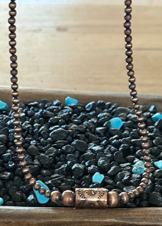 Rustic copper pearl necklace with stamped barrel focal bead displayed on black stones and turquoise nuggets