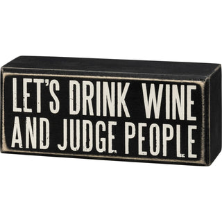A black box sign with white text that reads: LET'S DRINK WINE AND JUDGE PEOPLE.